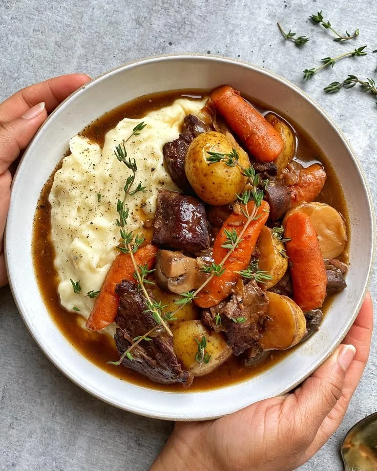 A bowl of red wine beef stew with potatoes and carrots served on a wooden table.